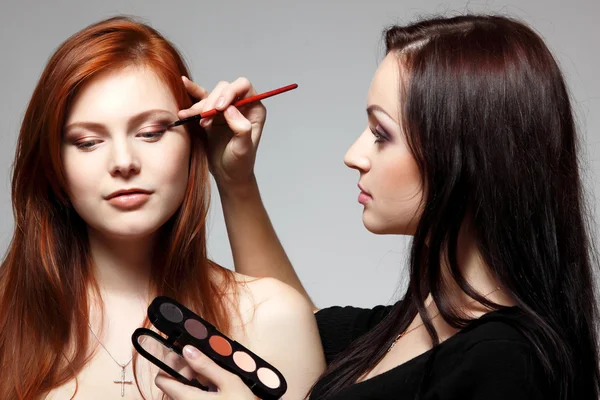 Retrato de una hermosa joven pelirroja con esteticista haciendo sombra de ojos de maquillaje —  Fotos de Stock