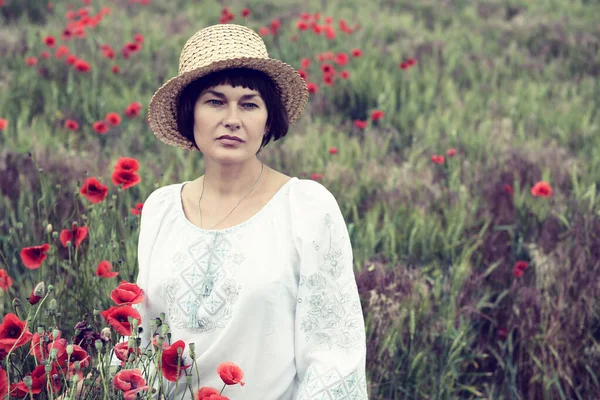 白いシャツに刺繍と藁帽子を身に着けている魅力的な中大人のウクライナ人女性はケシ畑 夏の屋外で歩く — ストック写真