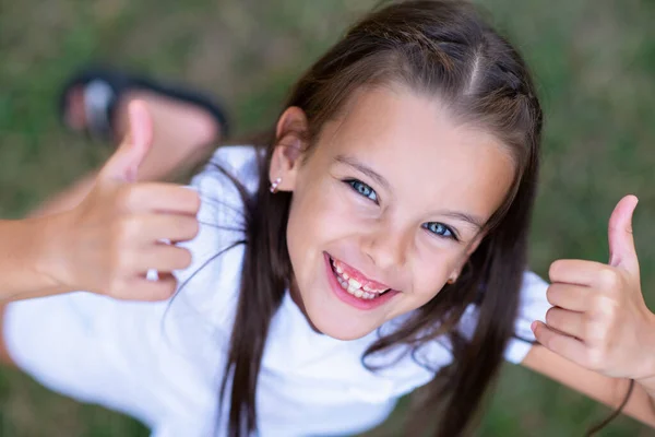 夏の自然の屋外で長い茶色の髪の親指を持つかわいい女の子 肯定的な表現で子供の肖像画 美しい笑顔の子供の顔 — ストック写真
