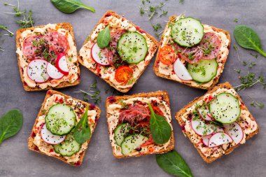 Krem peynirli, sebzeli ve salamlı sandviç. Salatalıklı sandviçler, turp, domates, gri arka planda salam, üst manzara. Düz yatıyordu.