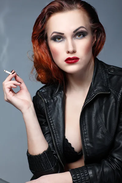 Ένα πορτρέτο του όμορφη σεξουαλική redheaded κορίτσι είναι σε ένα μαύρο δερμάτινο μπουφάν, καπνίζει ένα τσιγάρο — Φωτογραφία Αρχείου