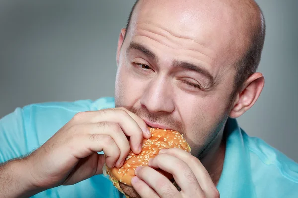 Homem engraçado comendo hambúrguer no fundo cinza — Fotografia de Stock
