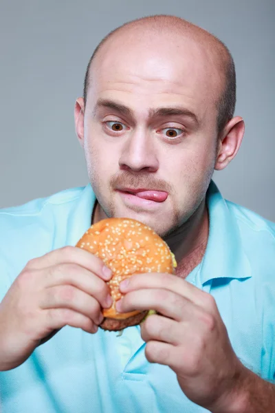 Uomo divertente mangiare hamburger su sfondo grigio — Foto Stock