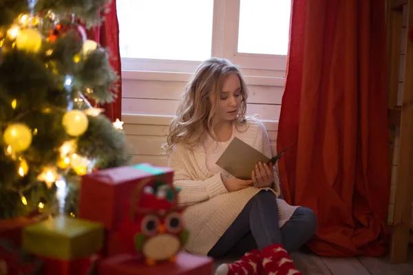 Giovane bella donna seduta sul pavimento vicino all'albero di Natale e leggere cartolina di Natale Immagine Stock