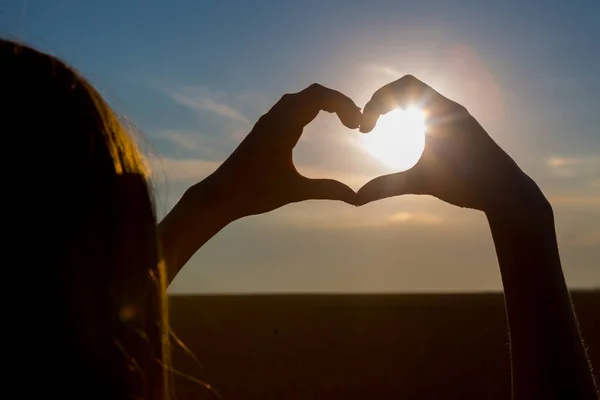 Fille faisant symbole de coeur avec ses mains au coucher du soleil sur fond de nature Image En Vente