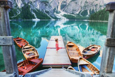 Kırmızı elbiseli ve şapkalı bir kız gölün ortasında teknelere ve dağlara tepeden bakan tek başına duruyor. Göl dağları yansıtır. Bries Gölü. Lago di Bries 