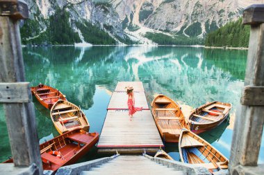 Kırmızı elbiseli ve şapkalı bir kız gölün ortasında teknelere ve dağlara tepeden bakan tek başına duruyor. Göl dağları yansıtır. Bries Gölü. Lago di Bries 