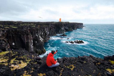 Kırmızı ceketli bir adam, uçurumun kenarında duran turuncu bir deniz fenerinin telefonunda fotoğraf çekiyor. İzlanda yaz gezisi 
