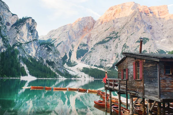 Kırmızı elbiseli ve kaslı şapkalı bir kız gölün ve dağların ortasındaki bir evin balkonunda oturuyor. Göl dağları yansıtır.