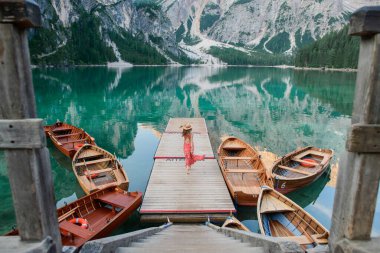 Kırmızı elbiseli ve şapkalı bir kız gölün ortasında teknelere ve dağlara tepeden bakan tek başına duruyor. Göl dağları yansıtır. Bries Gölü. Lago di Bries 