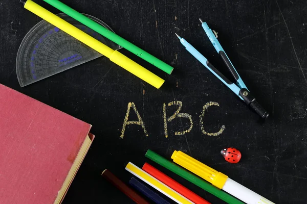 Канцелярские принадлежности (ручка, карандаш, линейка, компас) и книга на фоне черной школьной доски — стоковое фото