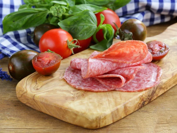 Копченая колбаса, помидоры и базилик на деревянной доске — стоковое фото