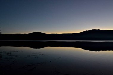 İspanya 'daki Sanabria Gölü' ndeki suyun üzerine yansıyan dağların siluetinin manzarası. Arka planda gün batımının turuncu ışığı.