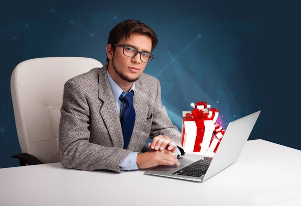 Hombre guapo sentado en el escritorio y escribiendo en el ordenador portátil con presente b —  Fotos de Stock