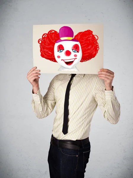 Businessman holding a cardboard with a clown on it in front of h — Stock Photo, Image