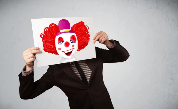 Businessman holding a cardboard with a clown on it in front of h — Stock Photo, Image