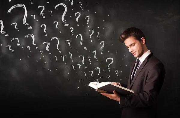 Young man reading a book with question marks coming out from it — Stock Photo, Image