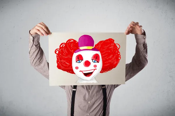 Businessman holding a cardboard with a clown on it in front of h — Stock Photo, Image