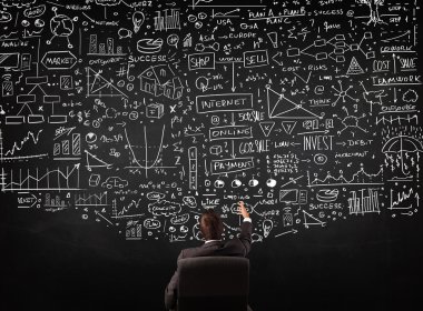 İşadamı elinde çizelgelerle bir tahtanın önünde oturuyor.