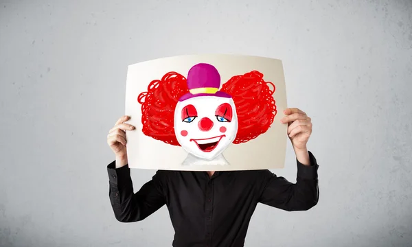 Businessman holding a cardboard with a clown on it in front of h — Stock Photo, Image