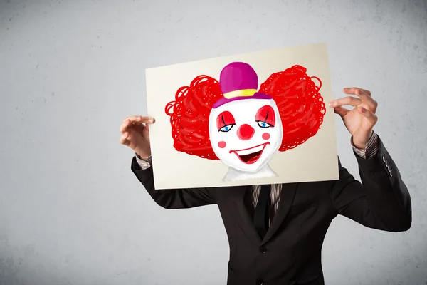 Businessman holding a cardboard with a clown on it in front of h — Stock Photo, Image