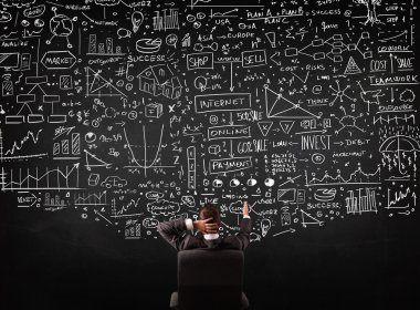 İşadamı elinde çizelgelerle bir tahtanın önünde oturuyor.