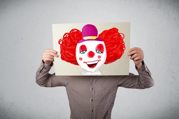 Businessman holding a cardboard with a clown on it in front of h — Stock Photo, Image