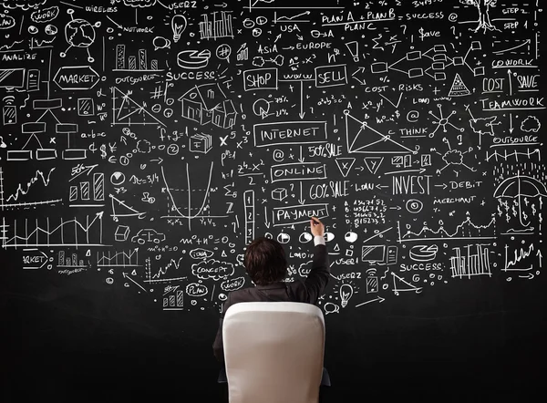 Businessman sitting in front of a blackboard with charts — Stock Photo, Image