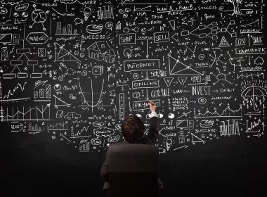 İşadamı elinde çizelgelerle bir tahtanın önünde oturuyor.