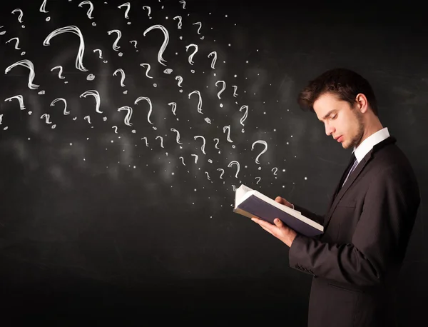 Young man reading a book with question marks coming out from it — Stock Photo, Image