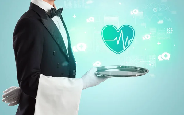 Przystojny młody kelner w smokingu trzymający ikony opieki zdrowotnej na tacy — Zdjęcie stockowe