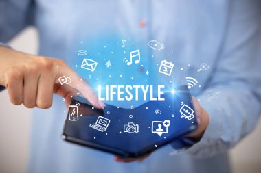 İş adamı, üzerinde LIFESTYLE yazıları ve sosyal medya kavramı olan katlanabilir bir akıllı telefon tutuyor.
