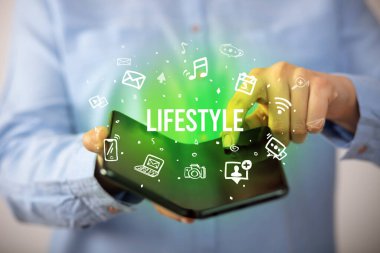 İş adamı, üzerinde LIFESTYLE yazıları ve sosyal medya kavramı olan katlanabilir bir akıllı telefon tutuyor.