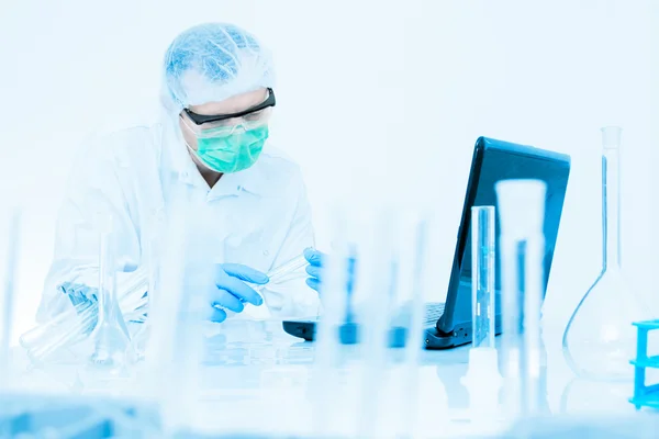 Científico trabajando en el laboratorio, en máscara protectora y tapa, examina un tubo de ensayo con líquido —  Fotos de Stock