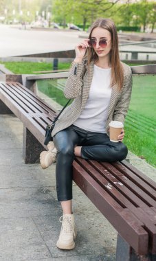 Kentteki bankta oturan bir fincan kahveyle güzel, genç moda tarzı bir kadın.
