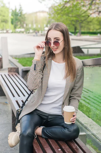 Kentteki bankta oturan bir fincan kahveyle güzel, genç moda tarzı bir kadın.