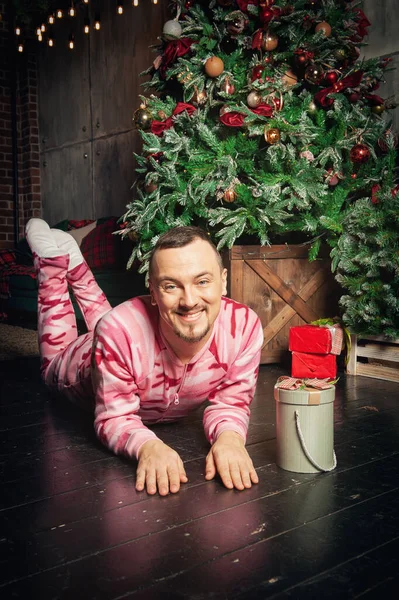 陽気面白いです男でピンクのスリープウェア装飾されたモミの木とクリスマスプレゼントの近くの床に横たわっ — ストック写真