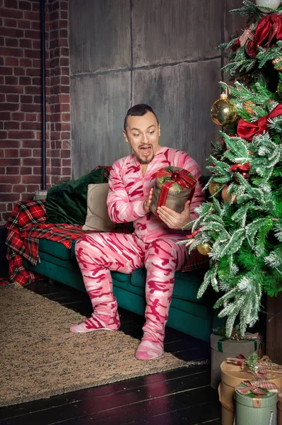 陽気な驚き面白い男でピンクの寝姿でソファの上に座っている装飾されたモミの木とクリスマスプレゼント — ストック写真