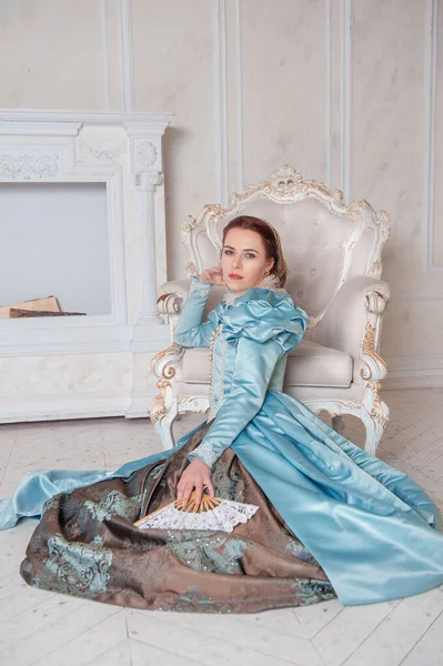 坐在扶手椅旁边的地板上 穿着中世纪风格的蓝色连衣裙的年轻貌美的女人 — 图库照片