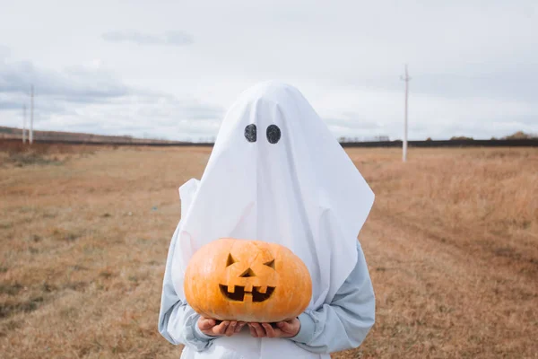 Dziecko w stroju ducha do halloween gospodarstwa rzeźbione dyni w jesiennym polu. — Zdjęcie stockowe