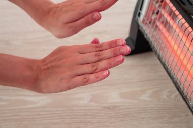 Kadın elektrikli ısıtıcının yanında ellerini ısıtıyor.