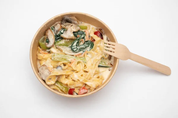 Pasta with vegetables in a cardboard lunch box with a wooden fork on a white background.