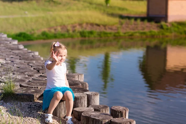 Cute little girl sitting near the lake waving to somebody — Stock Photo, Image