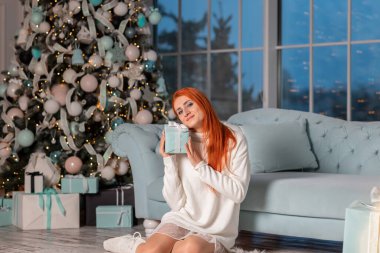 Güzel kızıl kız Noel arifesinde Noel ağacının yanında Noel hediyelerini açarken heyecanlanıyor.