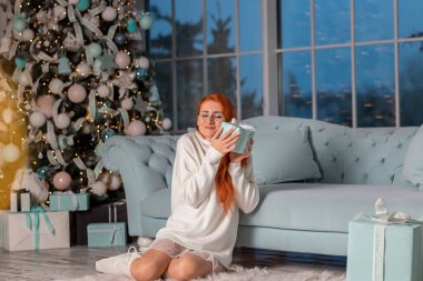 Güzel kızıl kız Noel arifesinde Noel ağacının yanında Noel hediyelerini açarken heyecanlanıyor.