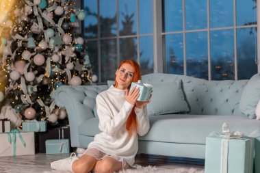 Güzel kızıl kız Noel arifesinde Noel ağacının yanında Noel hediyelerini açarken heyecanlanıyor.