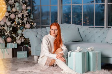 Güzel kızıl kız Noel arifesinde Noel ağacının yanında Noel hediyelerini açarken heyecanlanıyor.