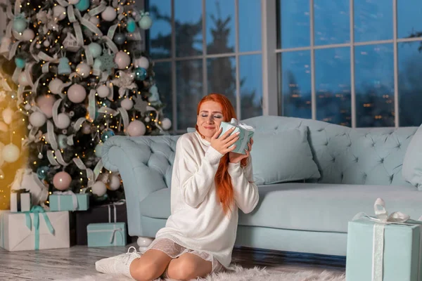 Güzel kızıl kız Noel arifesinde Noel ağacının yanında Noel hediyelerini açarken heyecanlanıyor.
