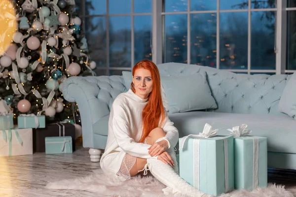 Güzel kızıl kız Noel arifesinde Noel ağacının yanında Noel hediyelerini açarken heyecanlanıyor.