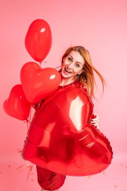 Kırmızı kalpli balon pozu veren kızıl saçlı güzel bir kız. Sevgililer günün kutlu olsun. Pembe arka planda dans eden güzel kızıl kızın stüdyo fotoğrafı..